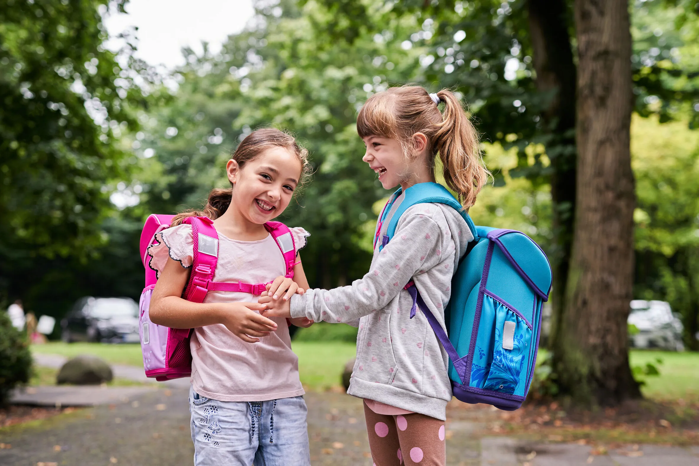 Zwei fröhliche Grundschulmädchen stehen mit ihren bunten Tornistern auf einem Weg im Grünen und lachen miteinander.