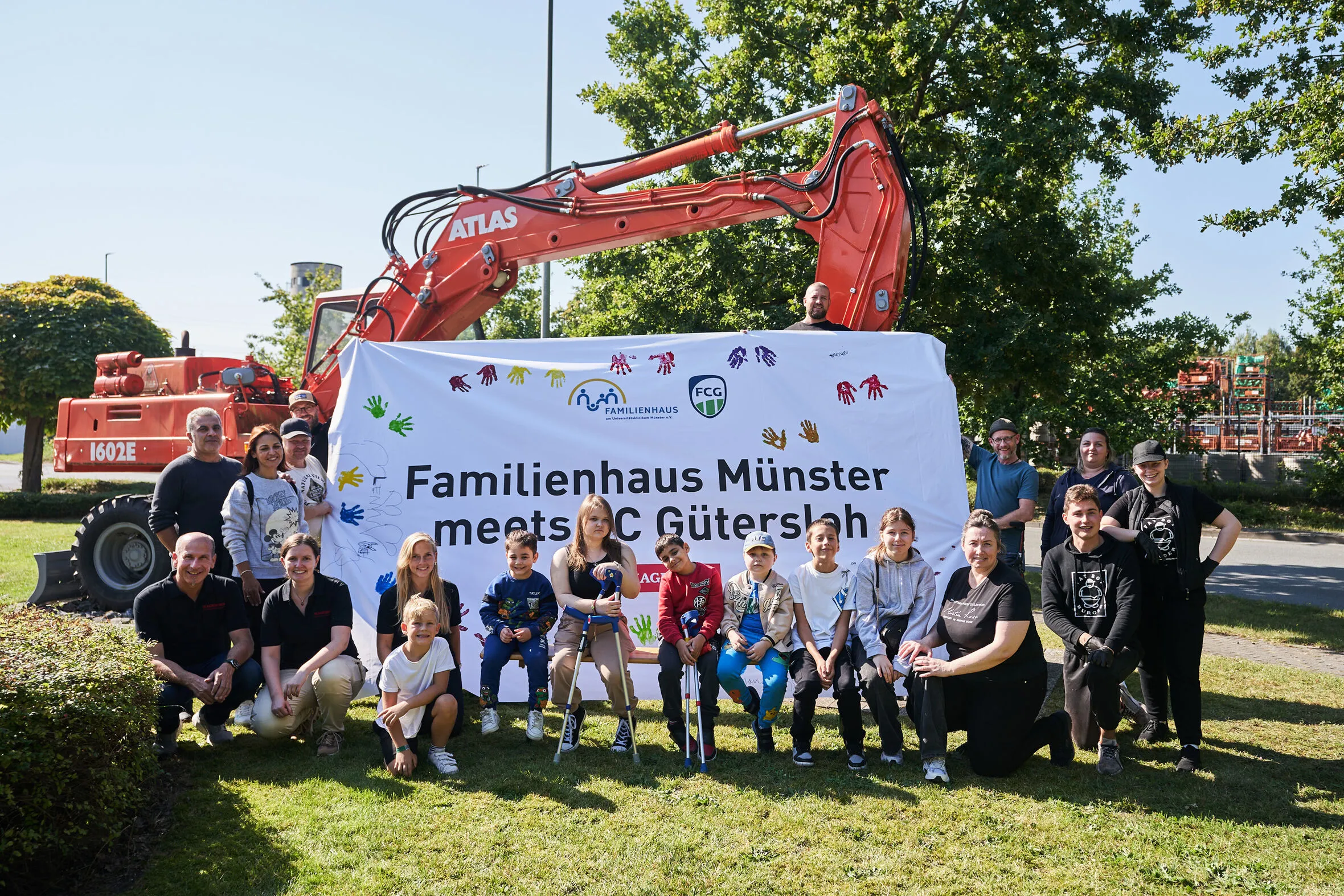 Kinder, Eltern, Helferinnen und Helfer lächeln gemeinsam in die Kamera. Gemeinsam präsentieren sie ihr selbst-gestaltetes Banner mit der Aufschrift „Familienhaus Münster meets FC Gütersloh“.