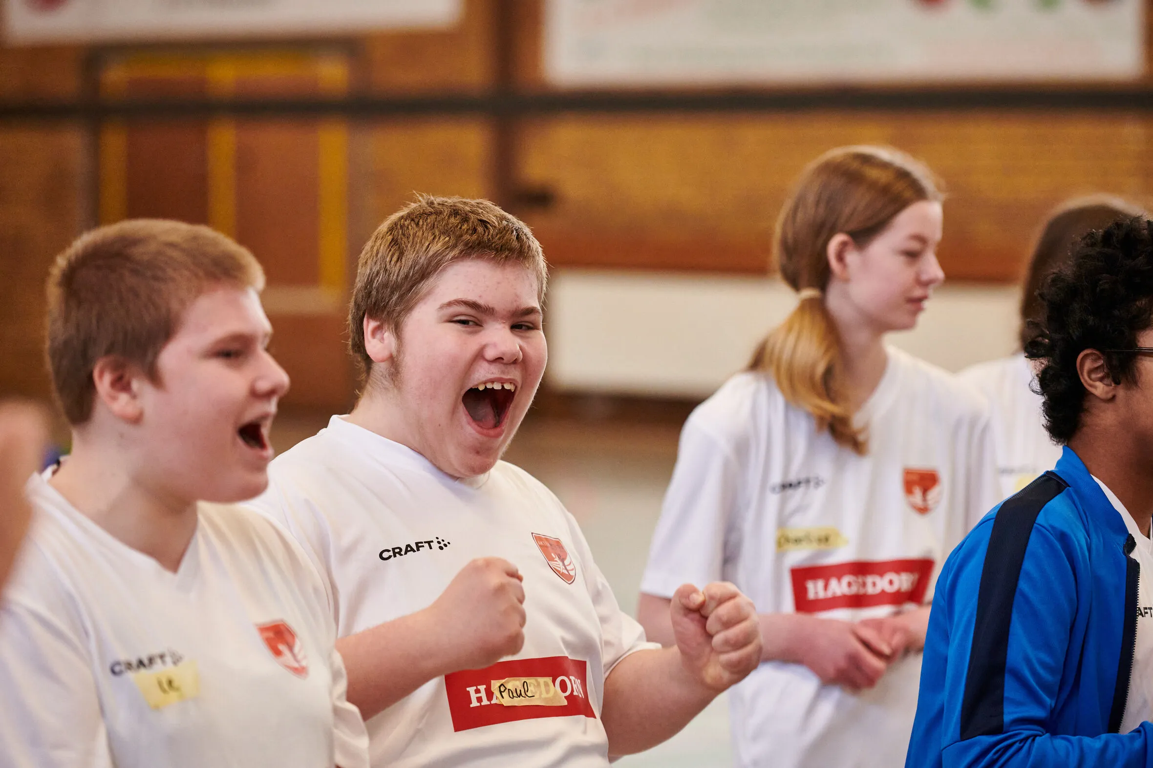 Jugendliche in weißen Trikots mit Hagedorn-Logo stehen in einer Sporthalle. Einer von ihnen zeigt mit einem breiten, offenen Lachen und geballten Fäusten große Freude und Begeisterung.