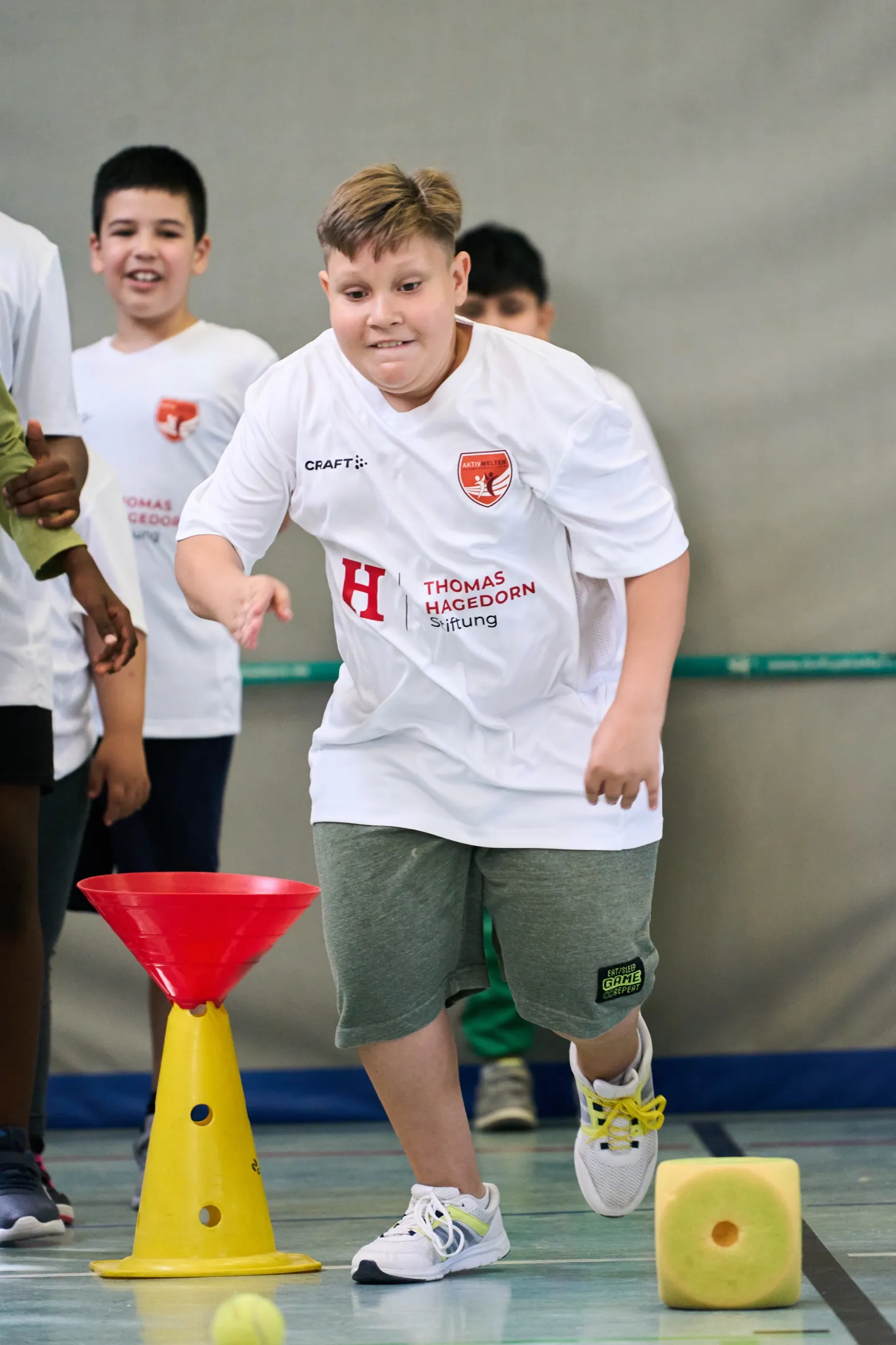Ein Junge im weißen Trikot der Thomas Hagedorn Stiftung läuft konzentriert durch einen Übungsparcours aus Hütchen, im Hintergrund feuert ihn ein lachender Mitschüler an.