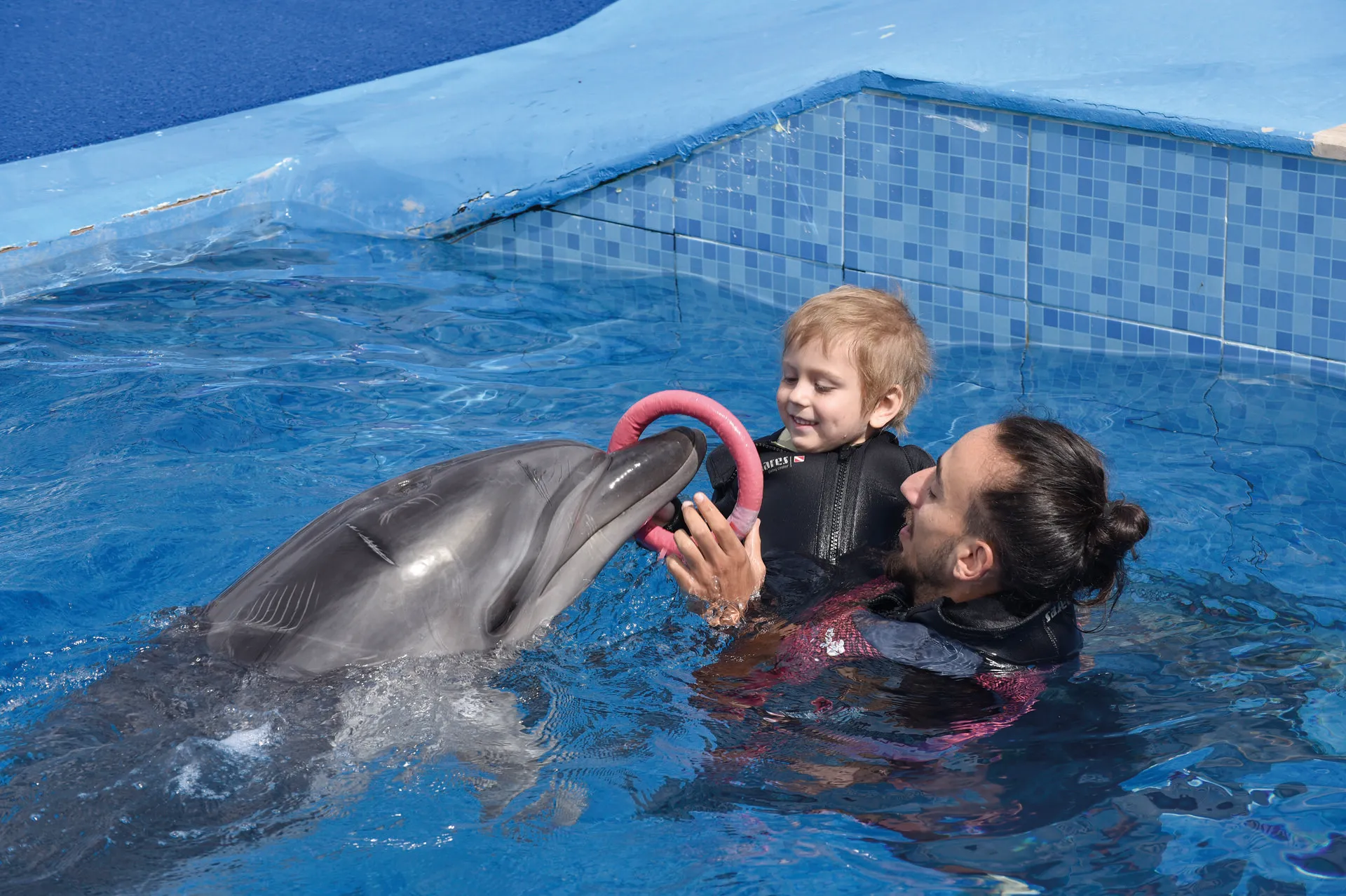 In den Armen eines Betreuers hängt ein Kind im Neoprenanzug einem Delfin im Wasser einen Schwimmring auf den Schnabel. Es macht den Anschein, als würden Kind und Delfin dabei lächeln.